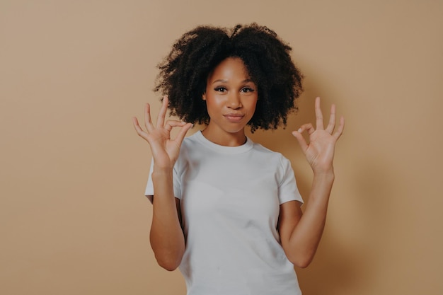 Junge afroamerikanische Dame hält beide Hände in einer „OK“-Geste isoliert vor braunem Hintergrund