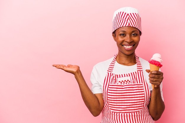 Junge afroamerikanische Eiscremeherstellerin, die Eis isoliert auf rosafarbenem Hintergrund hält und einen Kopienraum auf einer Handfläche zeigt und eine andere Hand an der Taille hält.