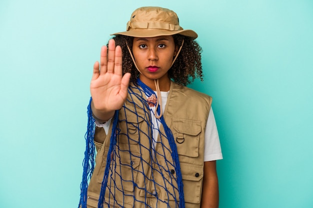 Junge afroamerikanische Fischerin, die ein Netz isoliert auf blauem Hintergrund hält, das mit ausgestreckter Hand steht und ein Stoppschild zeigt, um Sie zu verhindern.