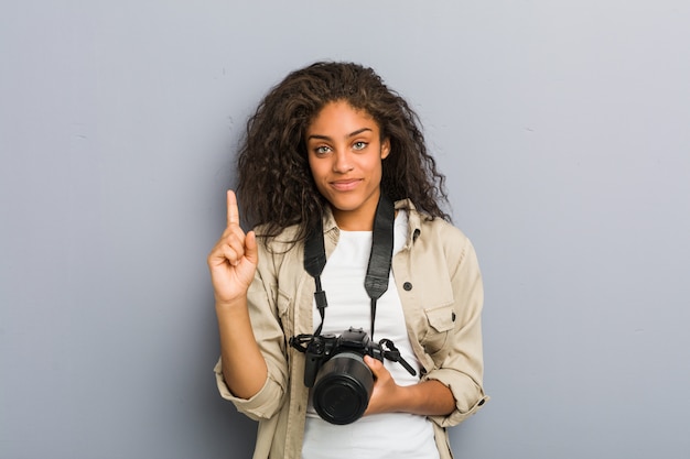 Junge afroamerikanische Fotograffrau, die eine Kamera hält, die Nummer eins mit Finger zeigt.