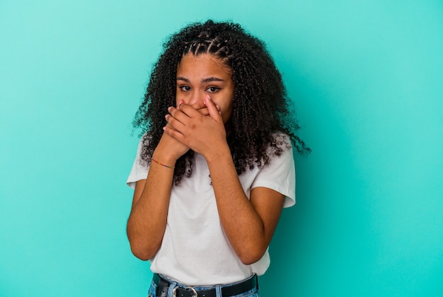 Junge afroamerikanische Frau auf blauem Hintergrund isoliert, die den Mund mit den Händen bedeckt, die besorgt aussehen.