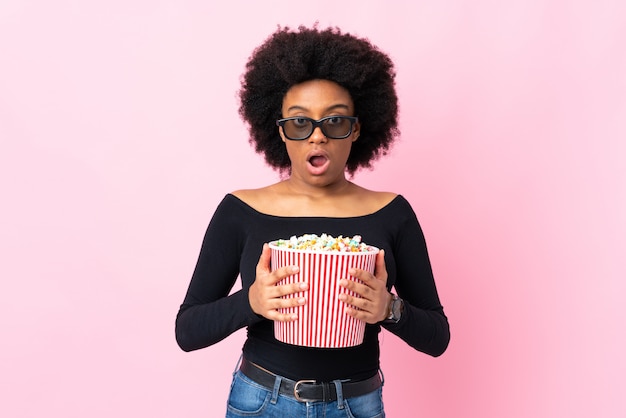 Junge afroamerikanische Frau auf rosa Wand überrascht mit 3D-Brille und hält einen großen Eimer Popcorn