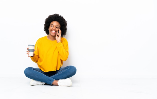 Junge afroamerikanische Frau, die auf dem Boden sitzt Kaffee hält, um und ein Handy wegzunehmen