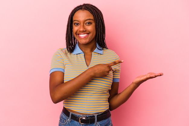 Junge afroamerikanische Frau, die auf rosafarbenem Hintergrund isoliert ist, aufgeregt, einen Kopienraum auf der Handfläche zu halten.