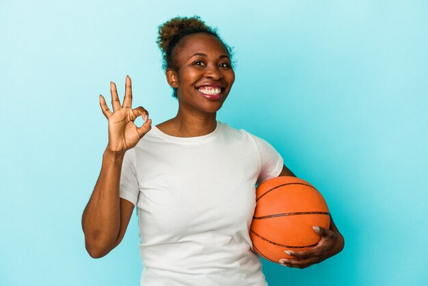 Junge afroamerikanische Frau, die Basketball isoliert auf blauem Hintergrund spielt, fröhlich und selbstbewusst, die ok Geste zeigt.