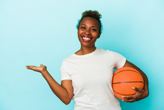 Junge afroamerikanische Frau, die Basketball spielt, isoliert auf blauem Hintergrund, der einen Kopienraum auf einer Handfläche zeigt und eine andere Hand an der Taille hält.