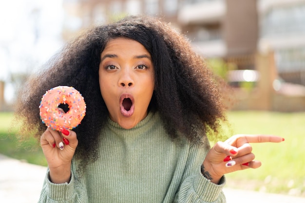 Foto junge afroamerikanische frau, die draußen einen donut hält, überrascht und auf die seite zeigt