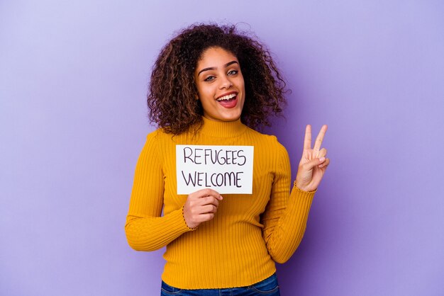 Junge afroamerikanische Frau, die ein Flüchtlings-Willkommensplakat hält, isoliert freudig und sorglos, ein Friedenssymbol mit den Fingern zeigend.