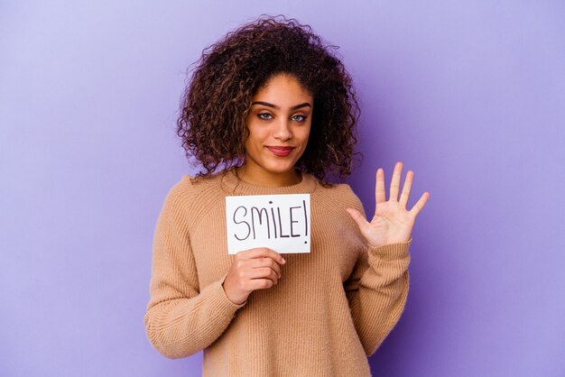 Foto junge afroamerikanische frau, die ein lächelnplakat auf lila lächelnd fröhlich zeigt nummer fünf mit den fingern hält.