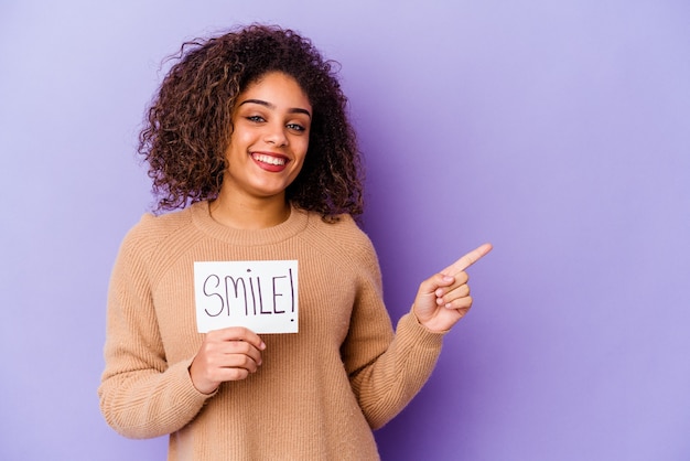 Junge afroamerikanische Frau, die ein Lächelnplakat lokalisiert auf der lila Wand hält, die lächelt und beiseite zeigt und etwas an der leeren Stelle zeigt