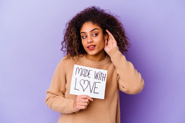 Junge afroamerikanische Frau, die ein Made with Love-Plakat hält, das auf lila Wand lokalisiert versucht, einen Klatsch zu hören