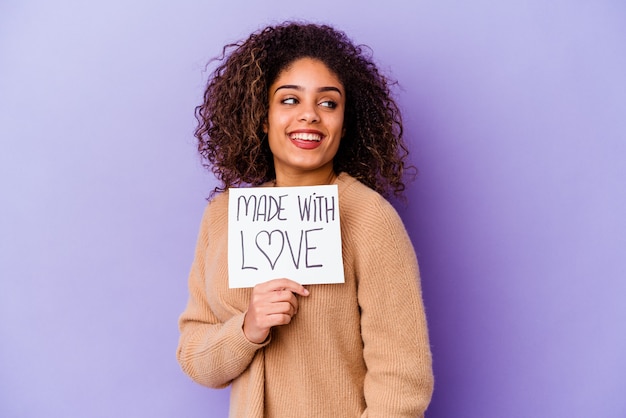 Junge afroamerikanische Frau, die ein mit Liebe hergestelltes Plakat hält, das auf lila lokalisiert aussieht, sieht lächelnd, fröhlich und angenehm beiseite.