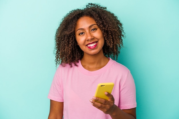 Junge afroamerikanische Frau, die ein Mobiltelefon auf blauem Hintergrund isoliert hält glücklich, lächelnd und fröhlich.