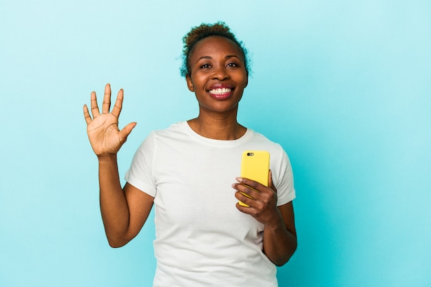 Junge afroamerikanische Frau, die ein Mobiltelefon auf blauem Hintergrund isoliert hält, lächelt fröhlich und zeigt Nummer fünf mit den Fingern.