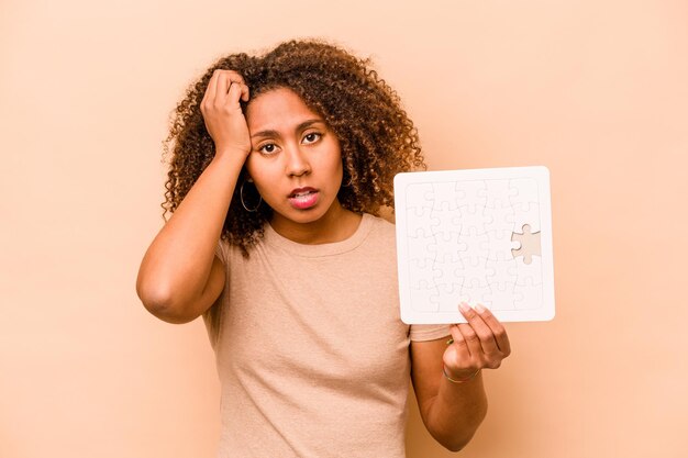 Junge afroamerikanische Frau, die ein Puzzle isoliert auf beigem Hintergrund hält, ist schockiert, dass sie sich an ein wichtiges Treffen erinnert hat