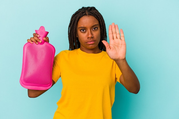 Junge afroamerikanische Frau, die eine heiße Tüte Wasser auf blauem Hintergrund isoliert hält, die mit ausgestreckter Hand steht und ein Stoppschild zeigt, um Sie zu verhindern.