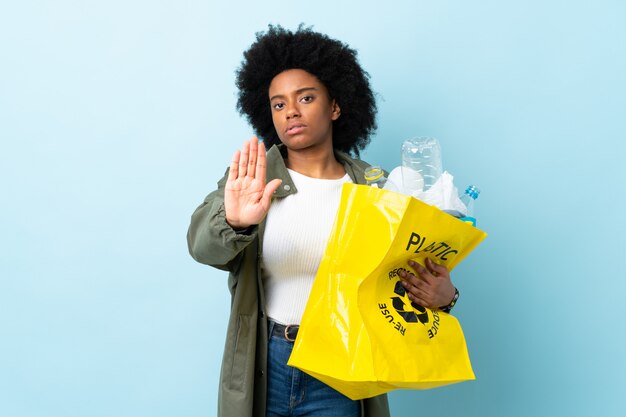 Junge afroamerikanische Frau, die eine Recycling-Tasche auf bunter Wand hält, die Stoppgeste macht