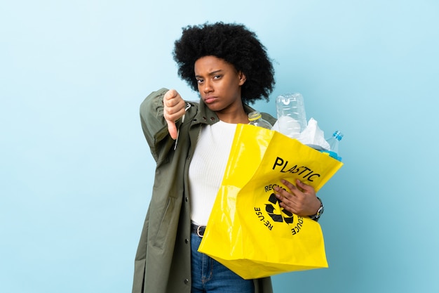 Junge afroamerikanische Frau, die eine Recycling-Tasche lokalisiert auf bunte Wand hält, die Daumen unten mit negativem Ausdruck zeigt