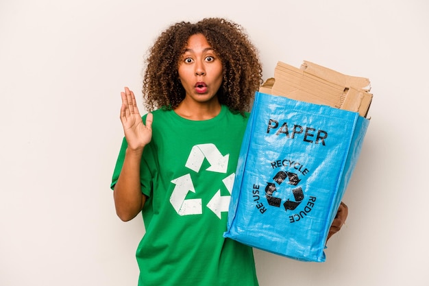 Junge afroamerikanische Frau, die einen Recycling-Beutel voller Papier hält, um isoliert auf weißem Hintergrund zu recyceln, überrascht und schockiert