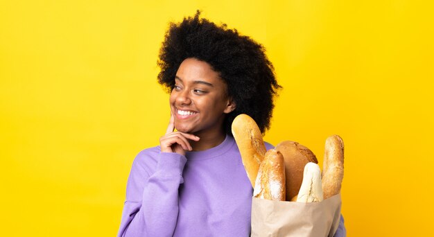 Junge afroamerikanische Frau, die etwas Brot kauft, das auf gelber Wand lokalisiert wird, die eine Idee denkt, während sie nach oben schaut