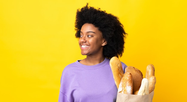 Junge afroamerikanische Frau, die etwas Brot lokalisiert auf gelbem Hintergrund kauft, das zur Seite schaut und lächelt