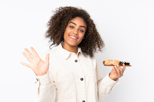Junge afroamerikanische Frau, die Sushi lokalisiert auf weißem Gruß mit Hand mit glücklichem Ausdruck hält