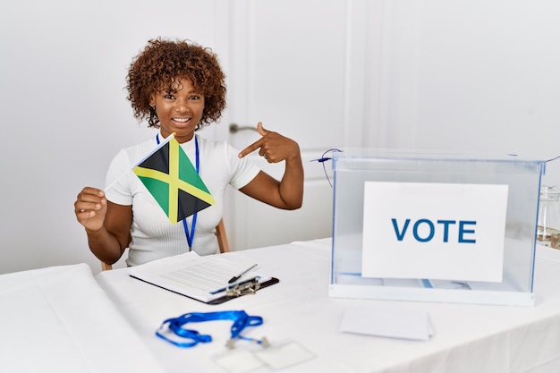 Junge afroamerikanische Frau im politischen Wahlkampf hält die Jamaika-Flagge in der Hand und zeigt mit dem Finger auf sich selbst, lächelt glücklich und stolz