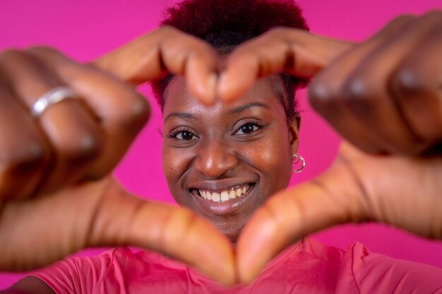 Junge afroamerikanische Frau isoliert auf rosa Hintergrund, lächelnd und mit Herzgesten im Studio fotografiert