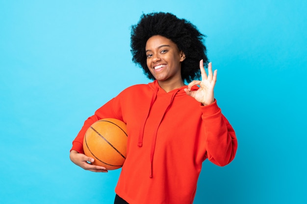 Junge afroamerikanische Frau lokalisiert auf Blau, das Basketball spielt und OK-Zeichen macht