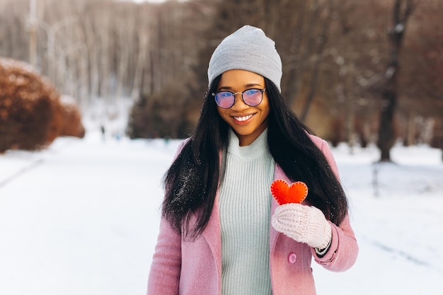 Junge afroamerikanische Frau mit Brille und warmen Handschuhen, die ein dekoratives rotes Herz halten