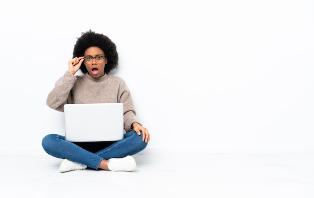 Junge afroamerikanische Frau mit einem Laptop, der auf dem Boden mit Brille sitzt und überrascht