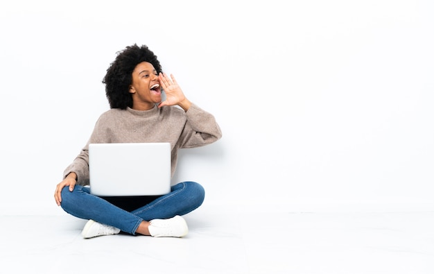 Junge afroamerikanische Frau mit einem Laptop, der auf dem Boden sitzt und mit weit offenem Mund zur Seite schreit