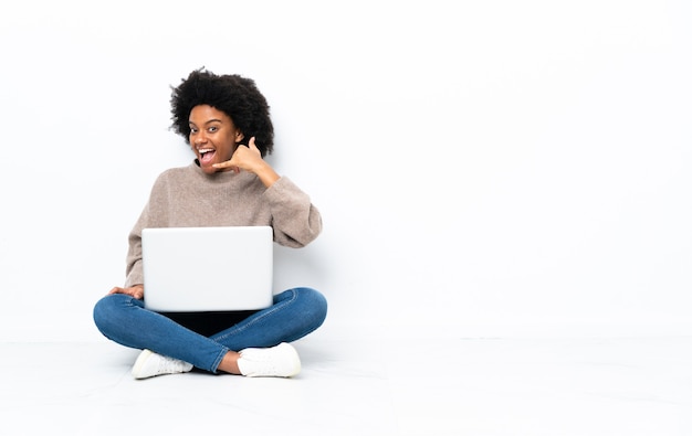 Junge afroamerikanische Frau mit einem Laptop, der auf dem Boden sitzt und Telefongeste macht. Rufen Sie mich zurück Zeichen