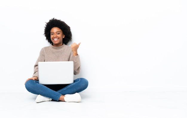 Junge afroamerikanische Frau mit einem Laptop, der auf dem Boden sitzt, zeigt zur Seite, um ein Produkt zu präsentieren