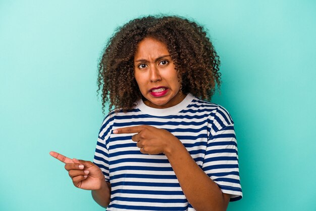 Junge afroamerikanische Frau mit lockigem Haar auf blauem Hintergrund isoliert schockiert und zeigt mit Zeigefingern auf einen Kopienraum.