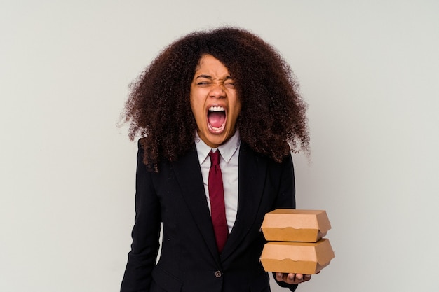 Junge afroamerikanische Geschäftsfrau, die einen Hamburger lokalisiert auf weißem Hintergrund hält, der sehr wütend und aggressiv schreit.