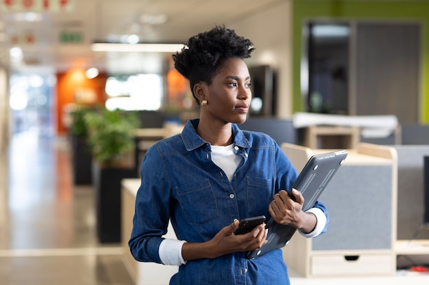 Foto junge afroamerikanische geschäftsfrau hält smartphone und laptop in der hand und schaut am arbeitsplatz weg