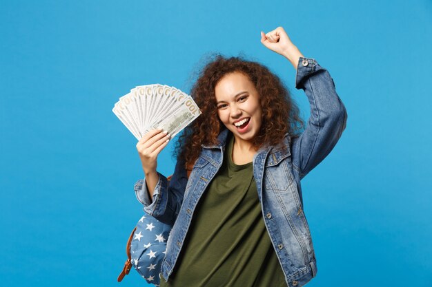 Junge afroamerikanische Mädchen Teen Studentin in Denim-Kleidung, Rucksack hält Bargeld isoliert auf blauer Wand cash