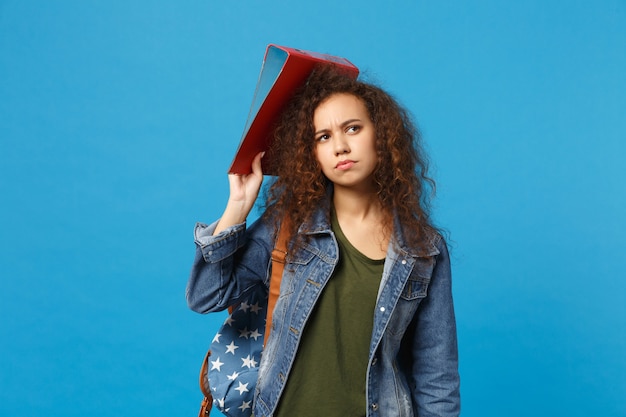 Junge afroamerikanische Mädchen Teen Studentin in Denim Kleidung Rucksack halten Ordner isoliert auf blauer Wand