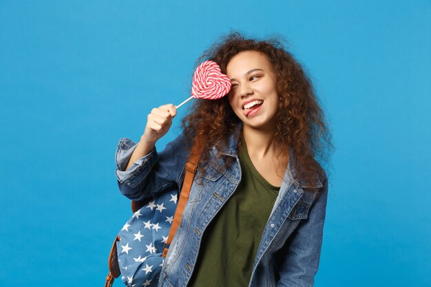 Junge afroamerikanische Mädchen Teen Studentin in Denim-Kleidung, Rucksack halten Süßigkeiten isoliert auf blauer Wand
