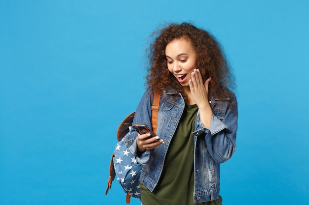 Junge afroamerikanische Mädchen Teen Studentin in Denim-Kleidung, Rucksack halten Telefon isoliert auf blauer Wand phone