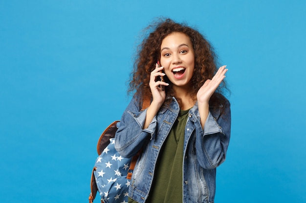 Junge afroamerikanische Mädchen Teen Studentin in Denim-Kleidung, Rucksack halten Telefon isoliert auf blauer Wand phone