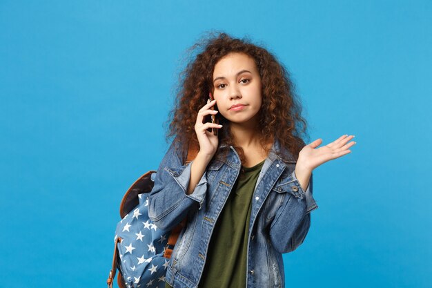 Junge afroamerikanische Mädchen Teen Studentin in Denim-Kleidung, Rucksack halten Telefon isoliert auf blauer Wand phone