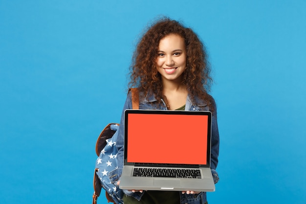 Junge afroamerikanische Mädchen Teen Studentin in Denim-Kleidung, Rucksackarbeit am PC isoliert auf blauer Wand