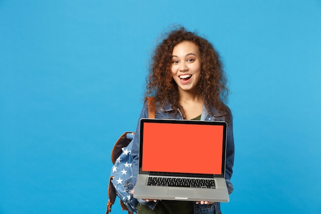 Junge afroamerikanische Mädchen Teen Studentin in Denim-Kleidung, Rucksackarbeit am PC isoliert auf blauer Wand