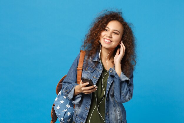 Junge afroamerikanische Mädchen-Teenager-Studentin in Denim-Kleidung, Rucksack-Kopfhörer isoliert auf blauer Wand