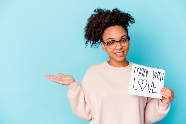 Junge afroamerikanische Mischrassenfrau, die ein mit Liebe gemachtes Konzept hält, das einen Kopienraum auf einer Handfläche zeigt und eine andere Hand auf Taille hält.