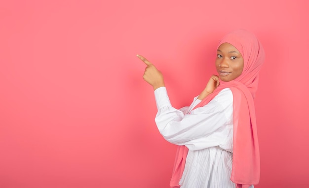 Foto junge afroamerikanische muslimische frau zeigt mit dem zeigefinger auf den freien raum, der in rosa schleier eingewickelt ist, isoliert auf rosafarbenem hintergrund.