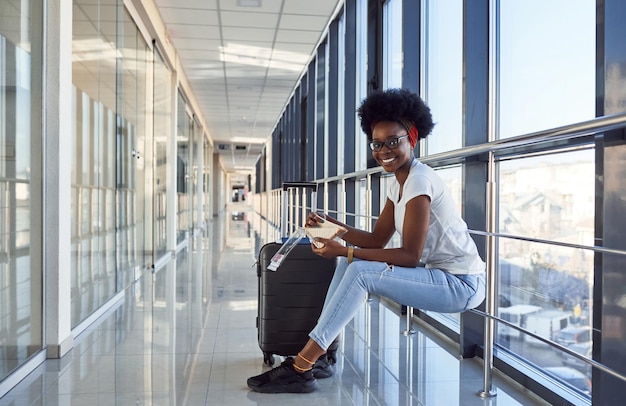 Junge afroamerikanische Passagierin in Freizeitkleidung ist am Flughafen mit Gepäck und isst etwas zu essen.