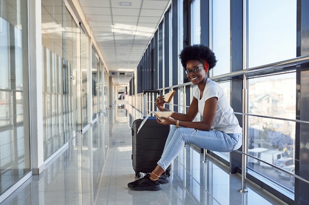 Junge afroamerikanische Passagierin in Freizeitkleidung ist am Flughafen mit Gepäck und isst etwas zu essen.